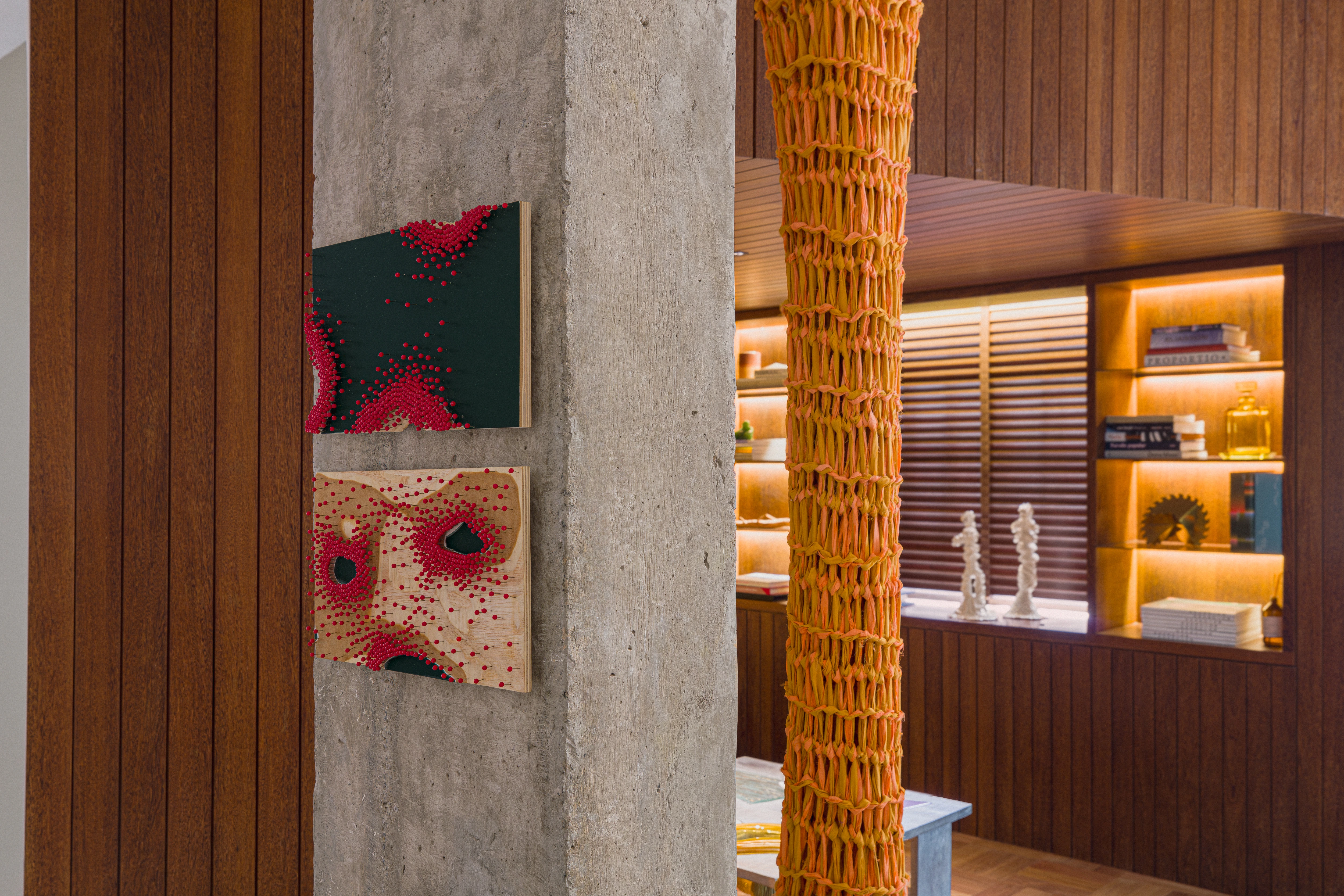 Detail of a concrete column with relief artwork and textile sculpture, next to a lit bookshelf with decorative objects and books.